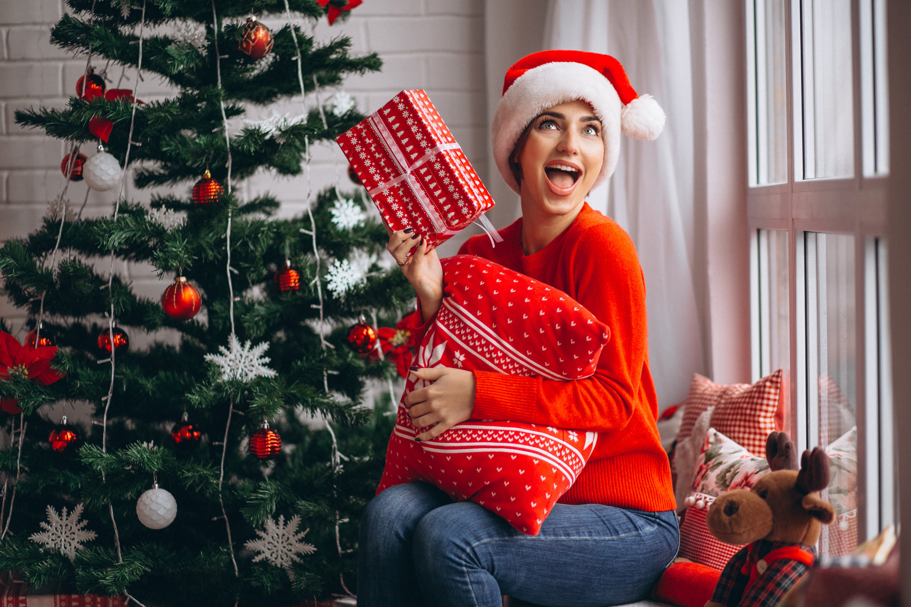Woman in red sweater and Santa hat holding a gift in front of a Christmas tree.